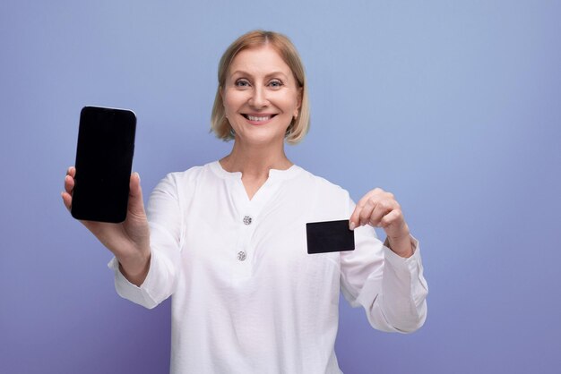 Mujer madura rubia delgada confiada en blusa blanca con tarjeta de plástico para ir de compras con maqueta