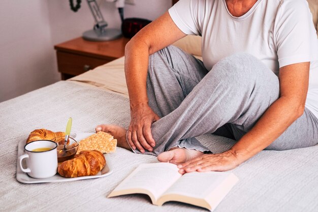 Mujer madura sentada en la cama leyendo un libro y comiendo croissants y creckers y bebiendo jugo de naranja - senior jubilado desayunando aislado y solo por la mañana