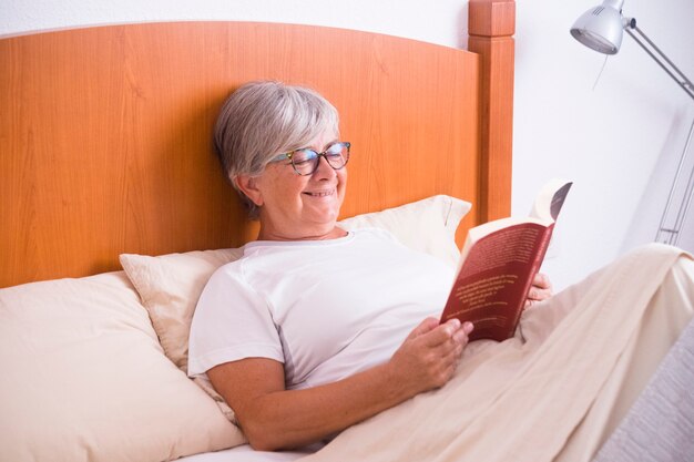 Mujer madura sentada en la cama leyendo un libro y sonriendo o riendo aislado y solo en la mañana