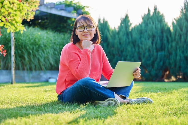 Mujer madura sentada en el césped verde en el patio trasero usando computadora portátil para ocio y trabajo mujer mirando la cámara trabajo remoto freelance relajación concepto de personas de mediana edad
