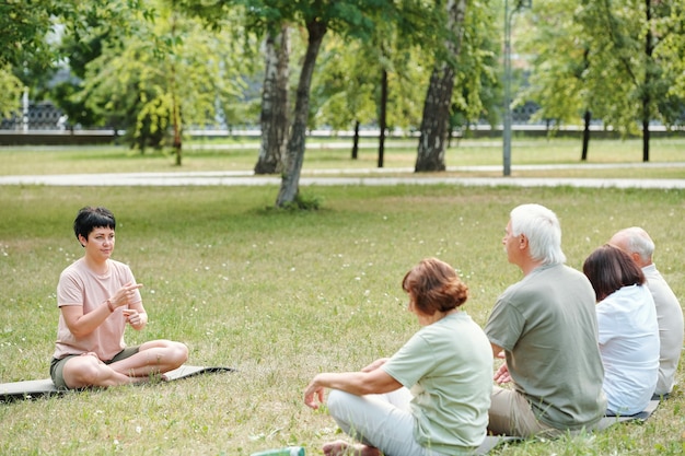 Mujer madura sentada sobre la hierba verde snd talknig a personas mayores durante las clases de terapia al aire libre en el parque