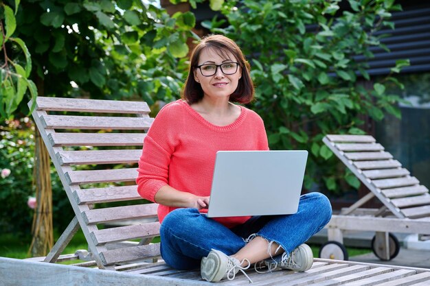 Mujer madura sentada en una tumbona de madera usando una computadora portátil para el ocio y el trabajo