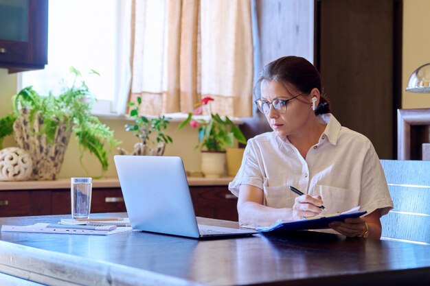 Mujer madura seria en auriculares hablando en video usando laptop