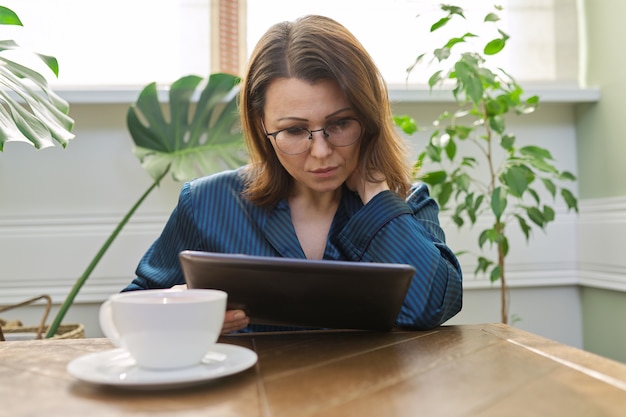 Mujer madura seria en casa leyendo correo, noticias, redes sociales en tableta digital. Mañana mujer en pijama con taza de té