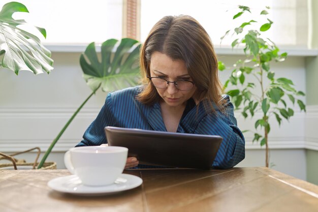 Mujer madura seria desayunando en casa en pijama con una taza de té sentada en la mesa leyendo una tableta digital. Mujer triste de leer correo, noticias. Emociones negativas, información, vida de mediana edad.