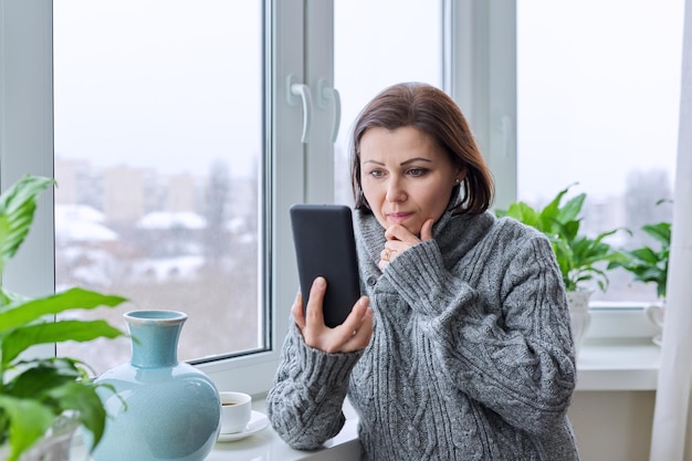 Mujer madura seria enfocada mirando la pantalla del teléfono inteligente Mujer de mediana edad en un suéter cálido en casa cerca de una ventana nevada de invierno