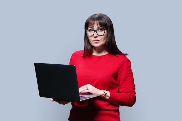 Mujer madura seria usando una computadora portátil mirando a la cámara en un fondo gris