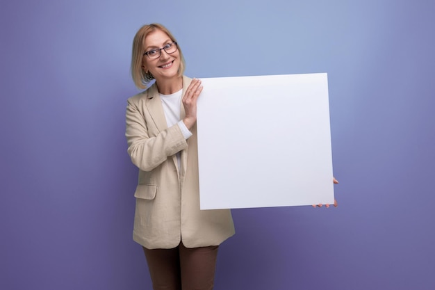 Mujer madura sonriente en chaqueta demostrando una maqueta de papel en blanco en el fondo del estudio con espacio de copia