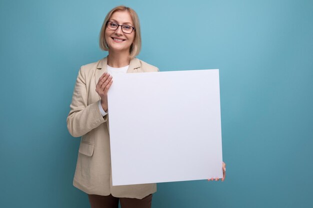 Mujer madura sonriente con chaqueta que muestra una maqueta de papel en blanco en el fondo del estudio con espacio para copiar