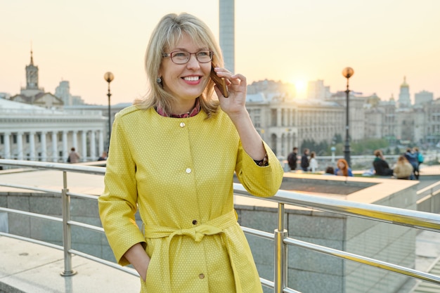 Mujer madura sonriente en gafas, abrigo amarillo hablando por teléfono móvil