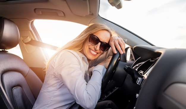 Mujer madura sonriente con gafas de sol y camisa azul sentada en el asiento del conductor dentro de un automóvil moderno y apoyando la cabeza en el volante Mujer de negocios haciendo parada durante la conducción
