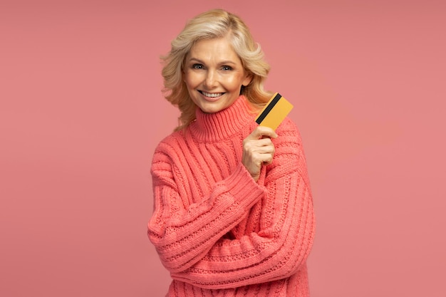 Mujer madura sonriente con suéter elegante con tarjeta de crédito en fondo rosa. compras, ventas