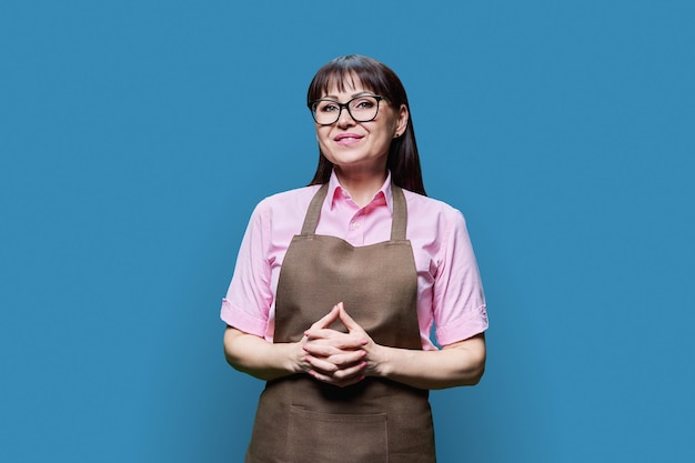 Mujer madura con una sonrisa positiva en delantal sobre un fondo azul de estudio