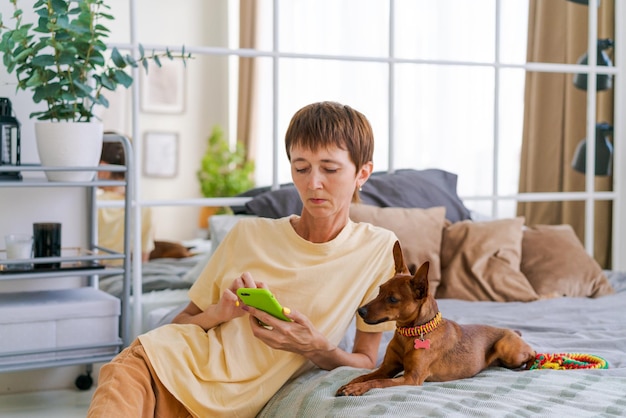 Mujer madura con un teléfono en la mano con un pinscher pigmeo en el dormitorio