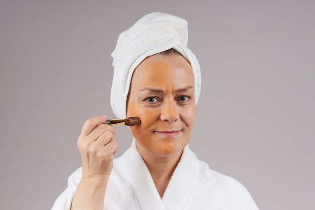 Una mujer madura con una túnica, una toalla blanca en la cabeza, se aplica una máscara de fruta naranja en la cara. Concepto de cuidado de la piel facial. sobre la pared gris.