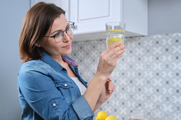 Foto mujer madura con un vaso de agua con limón en la cocina, antioxidante natural, alimentación saludable