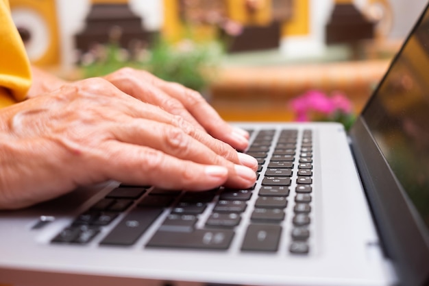 Mujer madura vestida de amarillo trabajando en una laptop desde casa con la mano en el primer plano del teclado