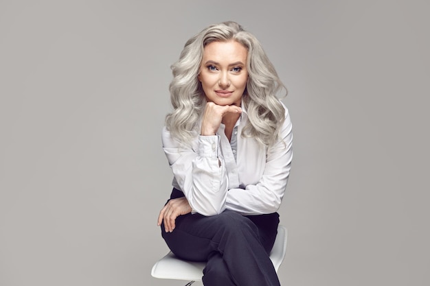 Mujer madura vestida con ropa de negocios casual elegante sentada en una silla sobre gris aislado