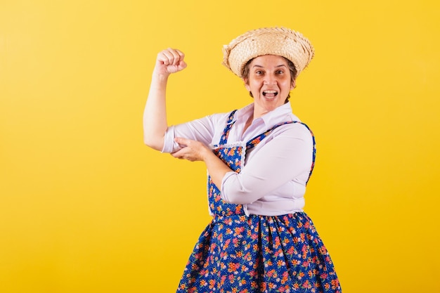 Mujer madura vestida con ropa típica de Festa Junina con la mano en el músculo bíceps ponderado sonriendo