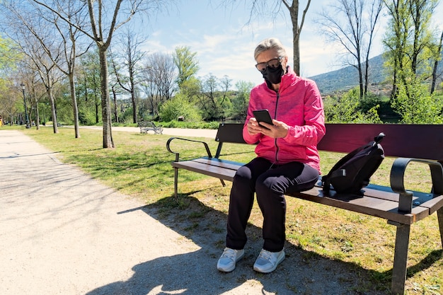 Mujer madura vistiendo máscara médica en un parque público comprobando su teléfono móvil sentado en un banco