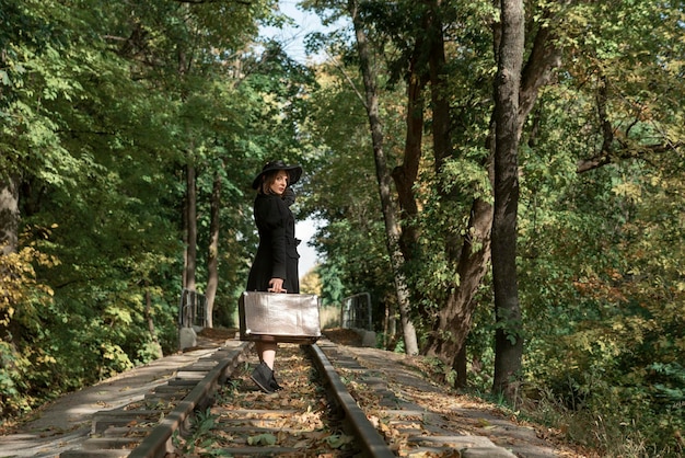 Mujer se va con una maleta a lo largo de los rieles a través del bosque de otoño Mujer vestida de negro Vista posterior