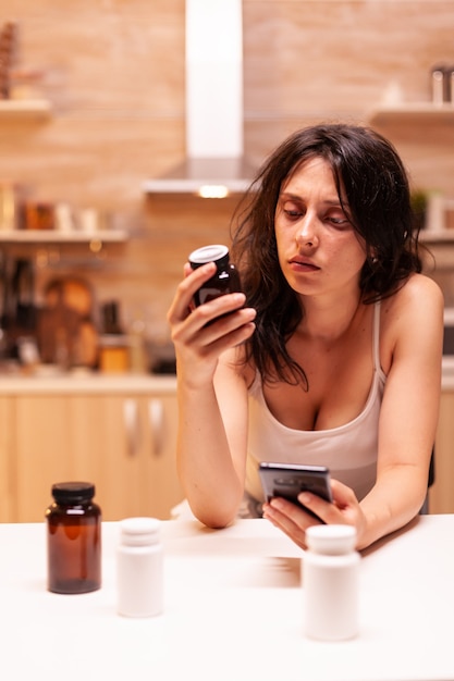 Mujer malsana que mira la botella de las píldoras que busca información en internet usando el teléfono. Esposa enferma preocupada que sufre de migraña, depresión, enfermedad y ansiedad sintiéndose agotada.