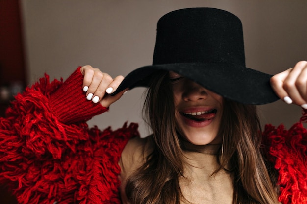 Mujer con manicura blanca vestida con suéter rojo se pone un sombrero negro y muestra su lengua