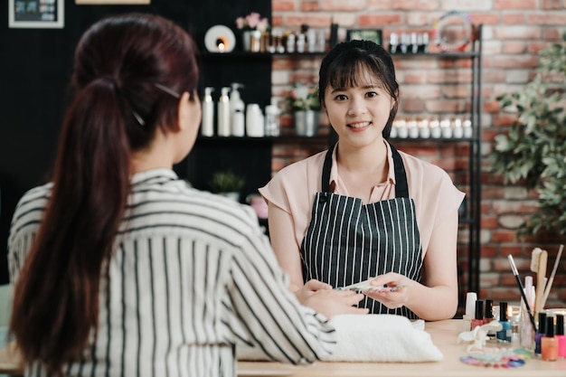 mujer manicurista dando manicura a una clienta asiática no reconocida en el salón de uñas. confiada joven esteticista cara cámara sonriendo en taller de belleza. elegante trabajador especialista en uñas en estudio.