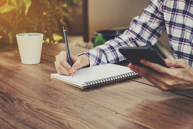 La mujer de la mano que escribe el cuaderno y que sostiene el teléfono en la tabla en jardín con el vintage entonó.