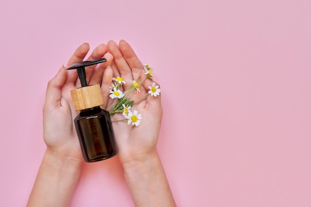 Mujer manos flores y botella de vidrio con producto cosmético sobre fondo rosa