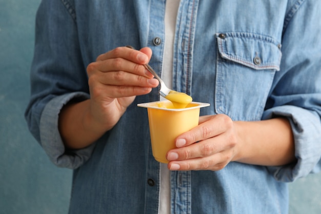 Mujer mantenga un vaso de plástico de yogur de crema agria y una cuchara contra el fondo azul.