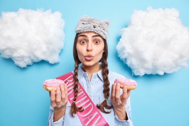 La mujer mantiene los labios doblados sostiene dos sabrosas rosquillas tiene la tentación de comer un postre dulce viste una camisa de dormir y una cinta de cumpleaños posa contra el azul