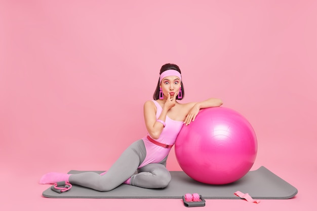 La mujer mantiene los labios doblados, se ve sorprendida por la cámara, usa maquillaje brillante, vestida con un mono, usa fitball para entrenar poses en la alfombra en toda su longitud.