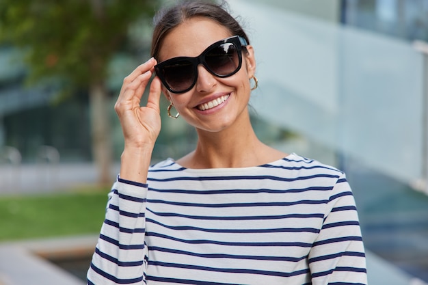 La mujer mantiene la mano en las gafas de sol sonríe agradablemente vestida con un jersey de rayas disfruta de tiempo libre día de verano tiene caminar poses al aire libre en borrosa