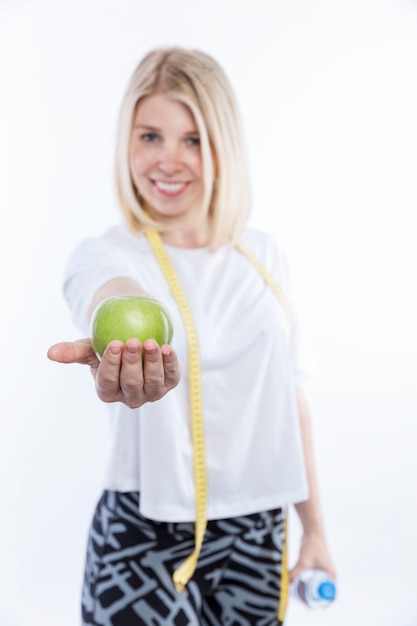 Mujer con una manzana verde y una botella de agua en sus manos Mujer rubia muy sonriente en una camiseta sin mangas blanca con una cinta métrica alrededor de su cuello Fondo blanco Vertical