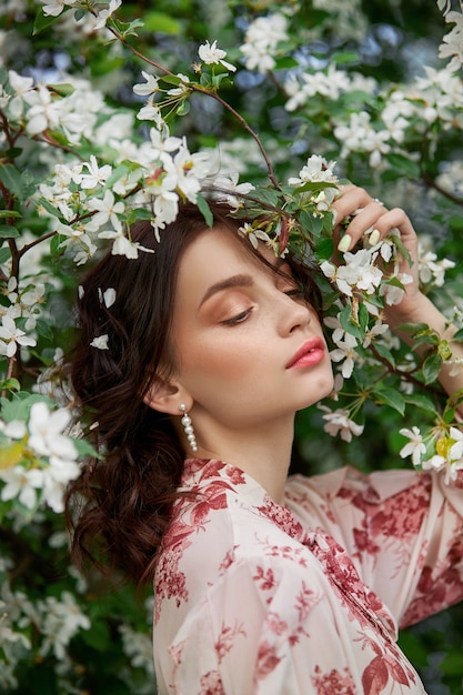 Mujer con maquillaje natural, cosméticos florales para la piel de la cara, niña posando en ramas de flores de manzano en flor en primavera