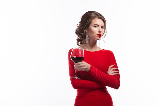 Mujer con maquillaje, peinado waering vestido rojo posando con copa de vid sobre fondo blanco, aislar.