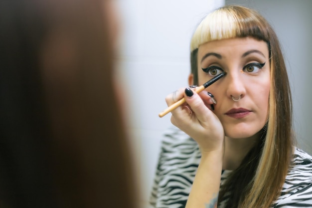 Mujer maquillándose frente al espejo del baño. Look casual con piercings y pelo teñido.