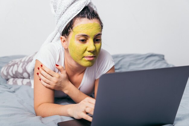 Foto mujer con máscara de belleza en la cara yacía en la cama con portátil