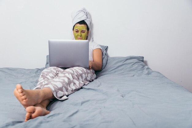 Mujer con máscara de belleza en la cara yacía en la cama con portátil