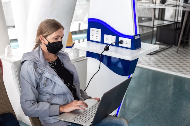 Una mujer con una máscara con una computadora portátil conectada a la red en el aeropuerto trabaja en línea Trabajo a distancia