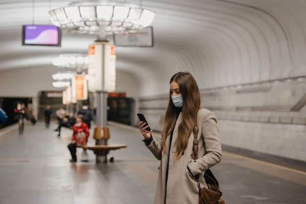 Una mujer con una máscara facial para evitar la propagación del coronavirus sostiene un teléfono inteligente en una estación de metro. Una niña con una mascarilla quirúrgica en la cara contra COVID-19 está esperando un tren en una plataforma de metro