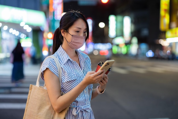 Mujer con máscara facial y uso de teléfono móvil en la ciudad por la noche