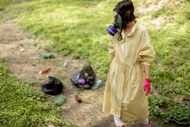 Mujer con máscara de gas y ropa protectora recogiendo basura plástica esparcida en el bosque