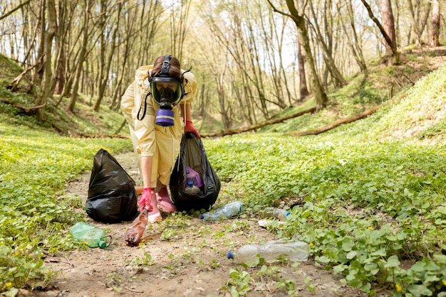 Mujer con máscara de gas y ropa protectora recogiendo basura plástica esparcida en el bosque