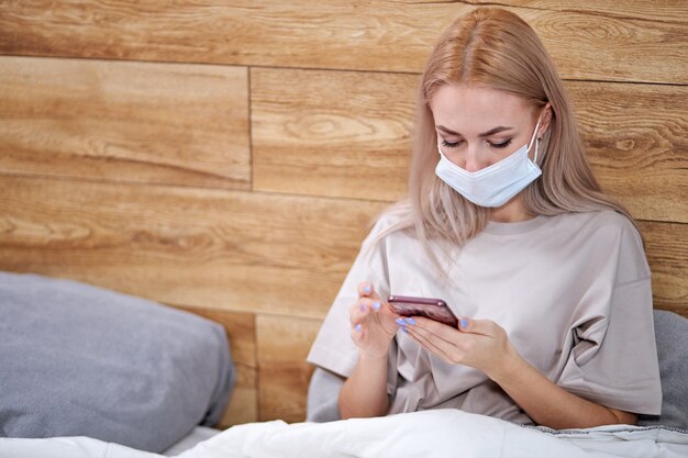 Mujer con máscara médica acostada en la cama con teléfono inteligente, descansando mientras está en cuarentena en autoaislamiento. resfriado, gripe, fiebre y migraña.