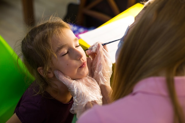 Una mujer con una máscara médica dibuja un patrón de aquagrim en la cara de un niño en el estudio frente a un espejo con lámparas. Diversión para los niños: colorear la cara. Rusia, Moscú, 15 de agosto de 2020