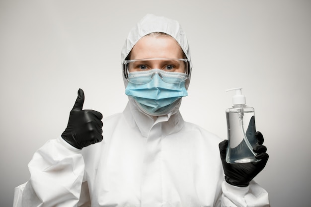 Mujer con máscara médica, gafas y traje protector blanco muestra la mano como signo.