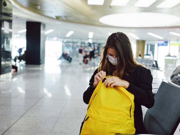 Mujer con máscara médica golpea la mochila amarilla del aeropuerto esperando