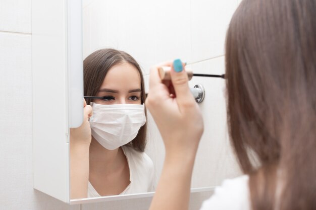 Mujer en máscara médica hace maquillaje, ojos de máscaras en casa en el baño